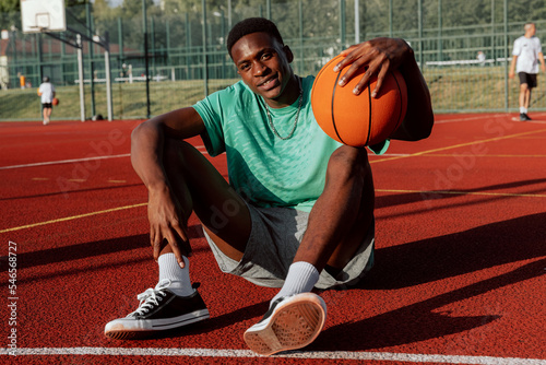 African american basketball player sitting on basketball court relaxing resting after exhausting training showing skills to friends comapny delighted man having break on background guys playing.
