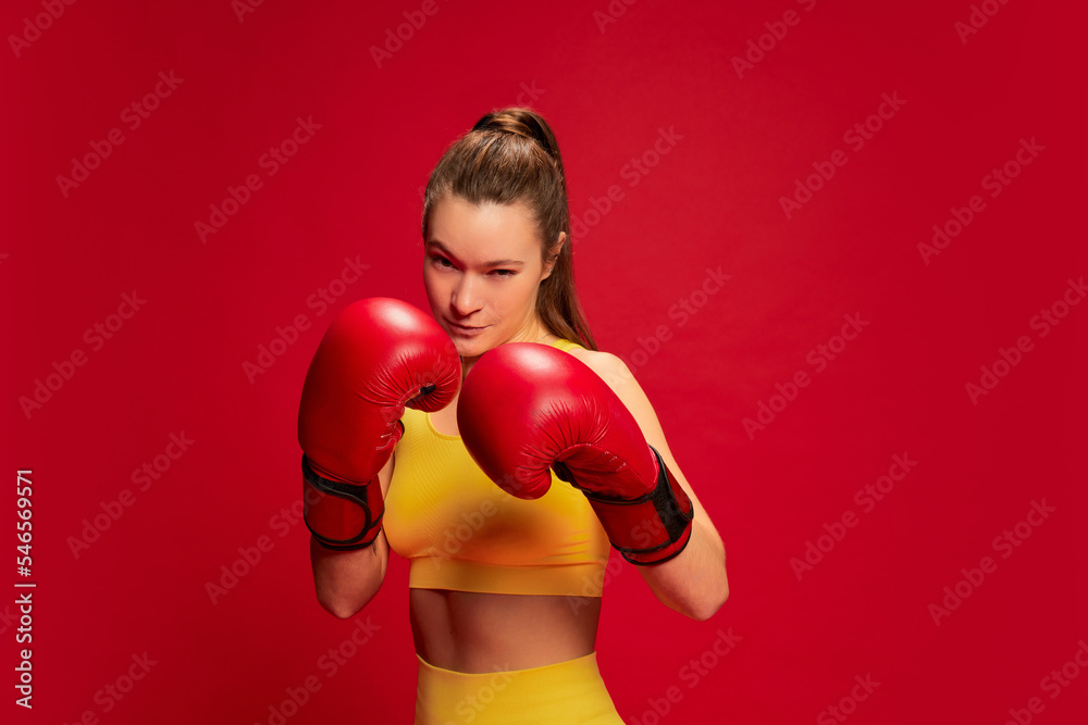 Portrait of young woman in yellow sportswear, boxing, training posing isolated over red background. Motivation