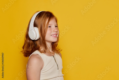 Portrait of little stylish girl with freckles wearing headphones isolated over bright yellow background. Concept of children emotions, dance, music, happiness