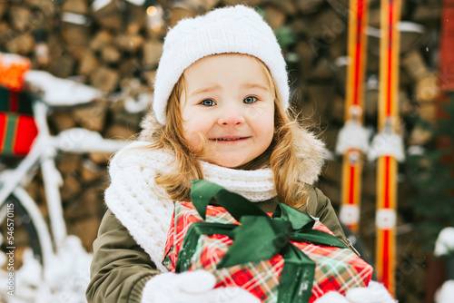 cute little girl in knitted white hat with gift present box stand at porch of country house decorared for celebration New Year and Christmas holidays for children outdoor photo