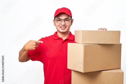 Cheerful smiling delivery guy in red shirt and cap standing and pointing piles of packages boxes