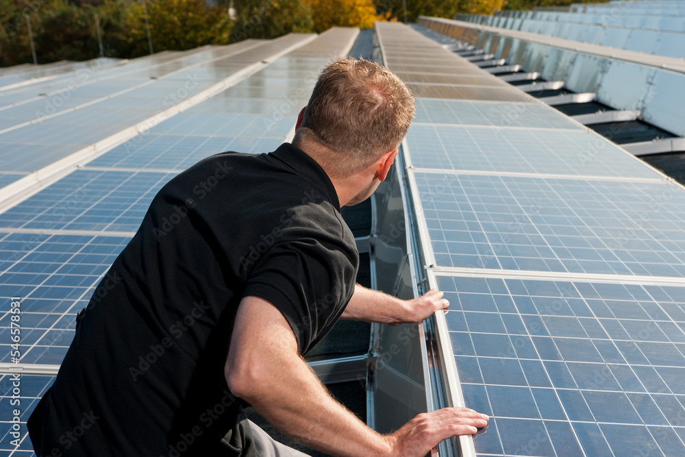 Looking over the shoulder of a workman checking a large array of solar panels on a roof in the warm summer light.