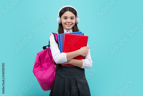 Back to school. Teenager school girl with backpack and headphones hold books ready to learn. School children on isolated blue studio background.