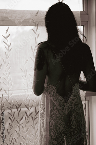 Underwear model in white lace slip dress, looking through window, upper body silhouette from the back