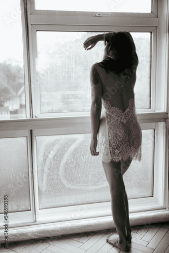 Underwear model silhouette in white lace slip dress looking through the window on a grey day