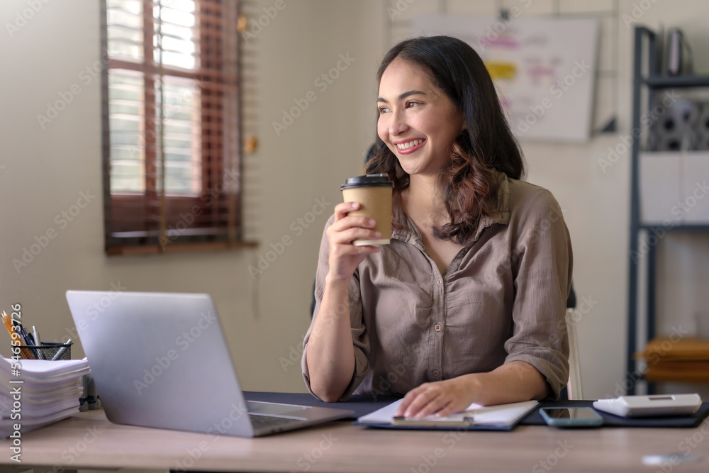 Beautiful Asian businesswoman sitting at her laptop happily working on her laptop and drinking coffee with a smile and looking at the camera looking bright in the office. Beautiful Asian businesswoman sitting at her laptop happily working on her laptop and drinking coffee with a smile and looking at the camera looking bright in the office.