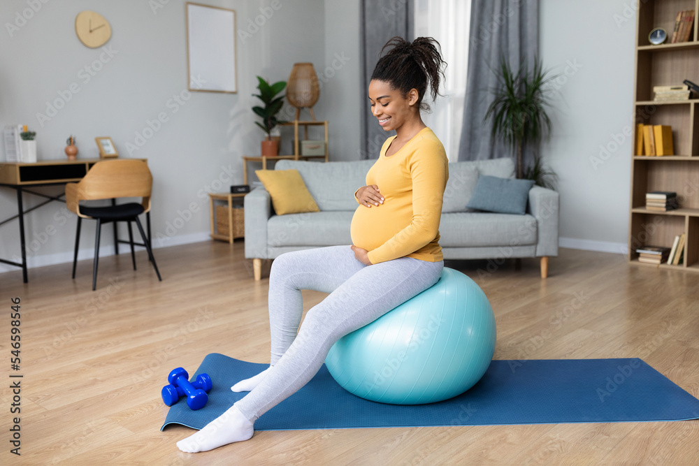 Smiling young black pregnant lady with big belly doing exercises on fitness ball and feels baby movements