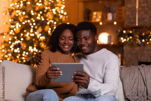 Smiling millennial black husband and wife have video call on tablet, watch video, sit on sofa in living room