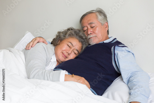 Asian elderly couple hugging and sleeping together on bed with white blanket, pillow in the bedroom at home. Retirement, health care, relax and spending time concept