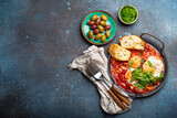 Middle Eastern and Maghrebi healthy dish Shakshouka made of eggs and tomato sauce served in pan with toasts, fresh cilantro and olives on rustic concrete background table from above, space for text
