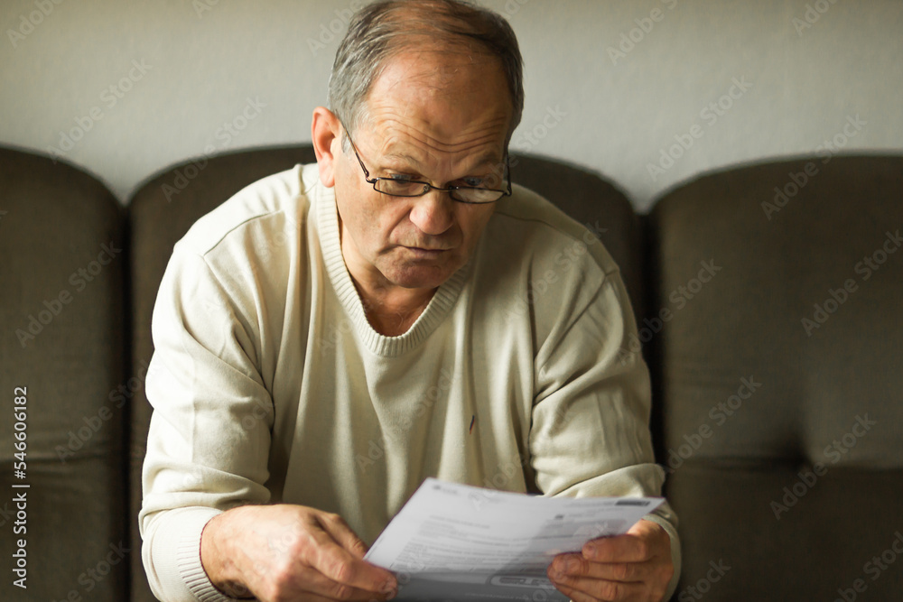 Fototapeta premium Worried senior man in white sweater and glasses reading utility bill in period of energy crisis. High heating prices.