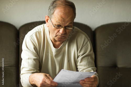 Worried senior man in white sweater and glasses reading utility bill in period of energy crisis. High heating prices.