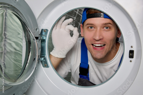 Cheerful worker in blue uniform shows hand gesture perfectly, looking out round hole washing machine layout. Experienced master advertises a company for installation and repair household appliances