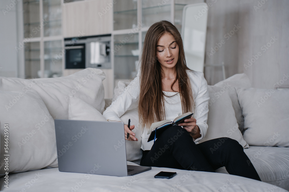 Beautiful young businesswoman in white jacket, black pants sitting on cozy sofa with laptop holds diary planning her day home. Italian student female preparing for distant lesson. Business and finance Beautiful young businesswoman in white jacket, black pants sitting on cozy sofa with laptop holds diary planning her day home. Italian student female preparing for distant lesson. Business and finance