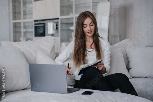 Beautiful young businesswoman in white jacket  black pants sitting on cozy sofa with laptop holds diary planning her day home. Italian student female preparing for distant lesson. Business and finance