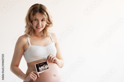 Image of pregnant smiling woman standing and posing while showing ultrasound scans over grey background . Looking at camera .