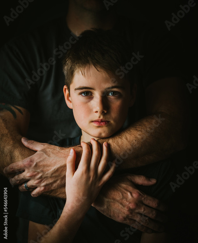 Close up portrait of serious boy with father's arms around him.