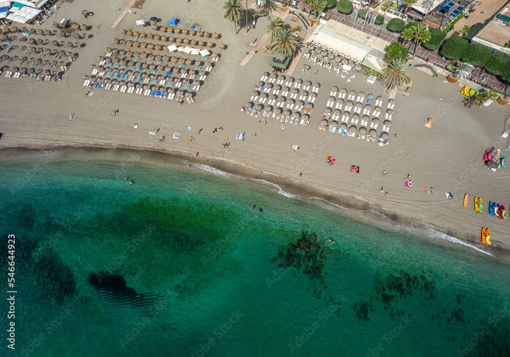 View from above, aerial view of an emerald and transparent Mediterranean sea with the beach full of beach umbrellas and tourists who relax and swim. Marbella, Spain.