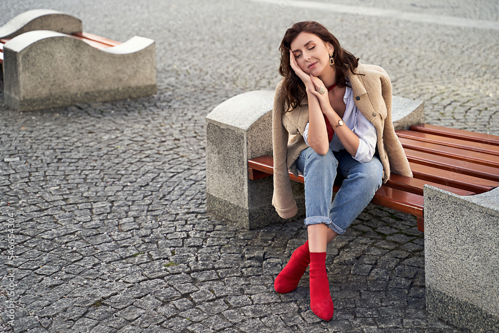 Beautiful young woman wearing teddy jacket jeans and white shirt sitting on a bench in the city