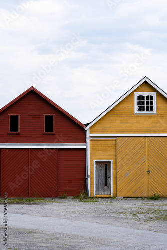 red and yellow fisherman's shed in Norway, which runs one over the other and has a beautiful view above it, but in the foreground there are some weeds growing between the gray gravel of the yard