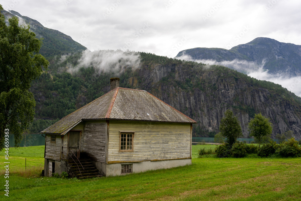 an old wooden house with a brown old tile roof, where green trees grow next to it, above which there is a fog and behind the fog are gray mountains