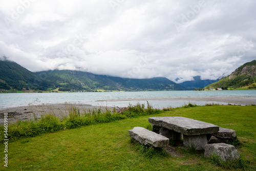 a stone table and benches stand in the grass by the blue fjord, which is a mountain caught in gray clouds