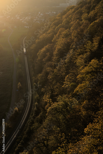A train travels on a curved railway track through a rural landscape of trees, forest, mountains and houses at sunset viewed from above from the air