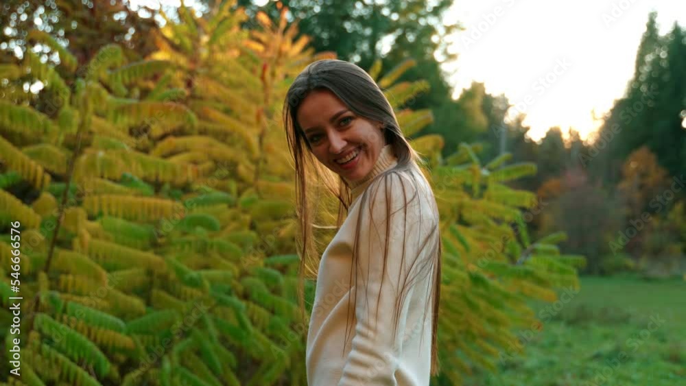 Happy smiling dark-haired woman waving her long hair in front of camera. Lady in white sweater posing at the backdrop of autumn park.
