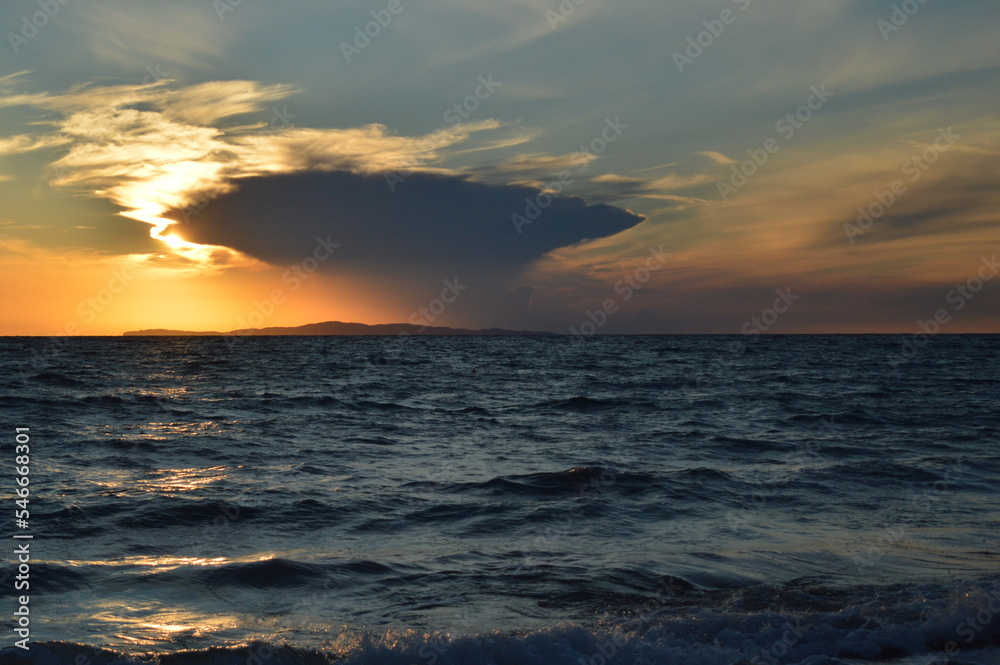 Obraz premium A dramatic sunset behind a small island in the distance with a large storm cloud above it