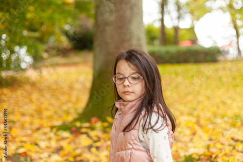 Little girl with long hair in glasses on a walk in the autumn park. Beautiful child in a warm vest outdoors