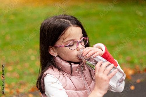 Cute girl with long hair and glasses drinks water from a plastic bottle on a blurred autumn park background