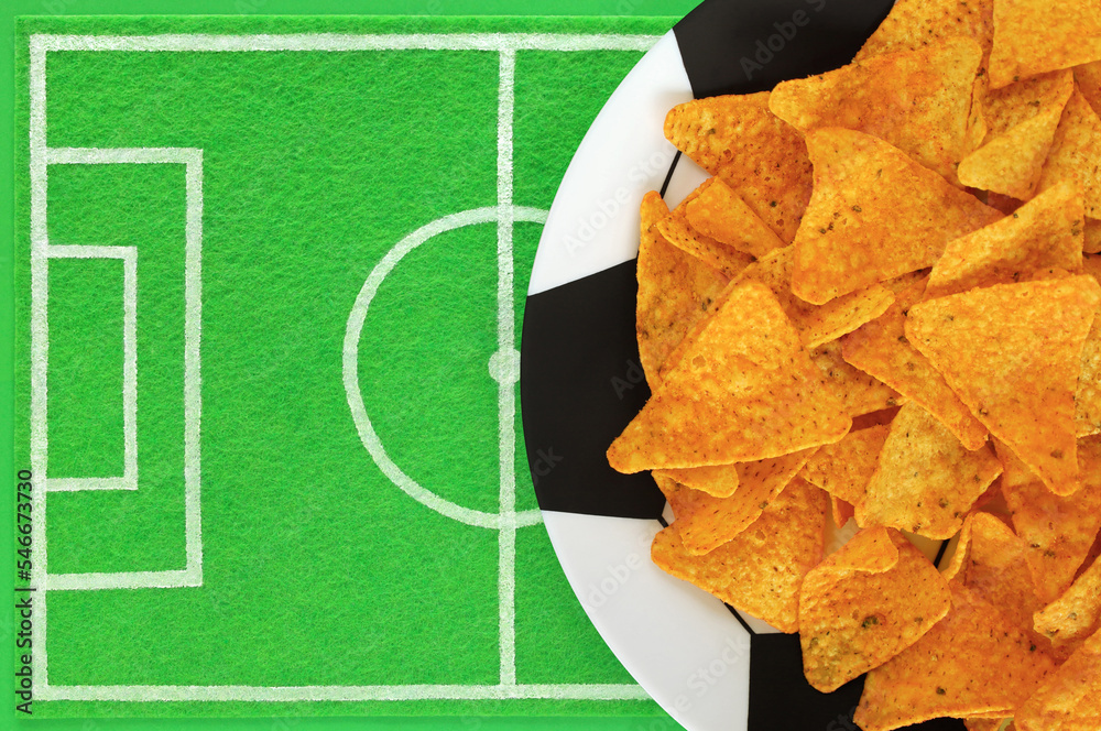 Mexican corn nacho chips on big plate painted like soccer ball, green felt dish mat like football field with place for text. Template of football soccer party table. Top view, copy space, mockup.