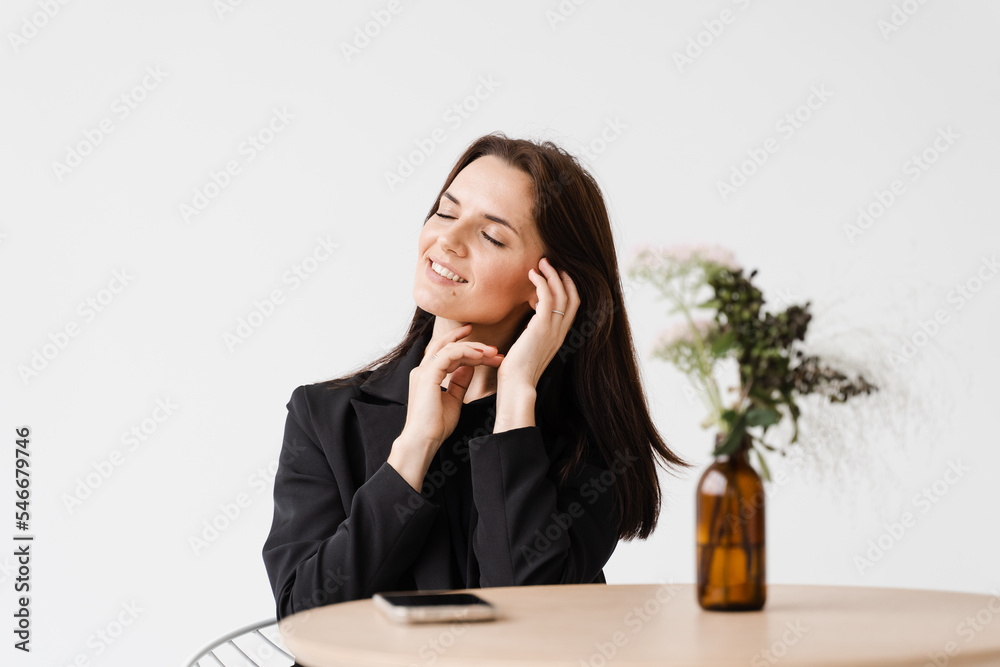 Dreamy cheerful girl dressed in business style is sitting at table in white cafe. Portrait of attractive young woman on white background. Dreamy cheerful girl dressed in business style is sitting at table in white cafe. Portrait of attractive young woman on white background.