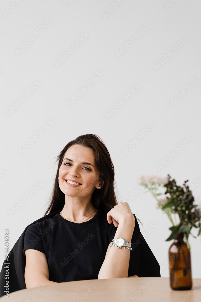 Happy confident woman is sitting at table on white background indoor in cafe. Smiling young woman dressed casual business style enjoy her lifestyle. Happy confident woman is sitting at table on white background indoor in cafe. Smiling young woman dressed casual business style enjoy her lifestyle.