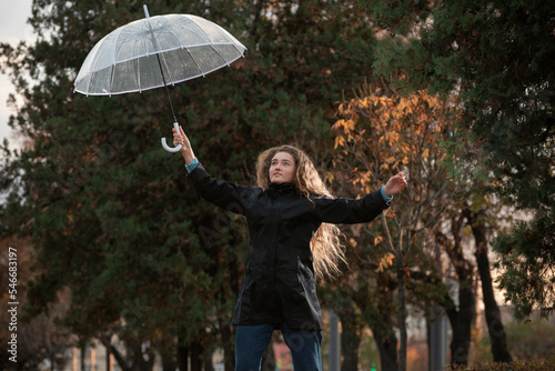 Smiling young student girl jumps on alley of autumn park with transparent umbrella. Lush and long hair of girl