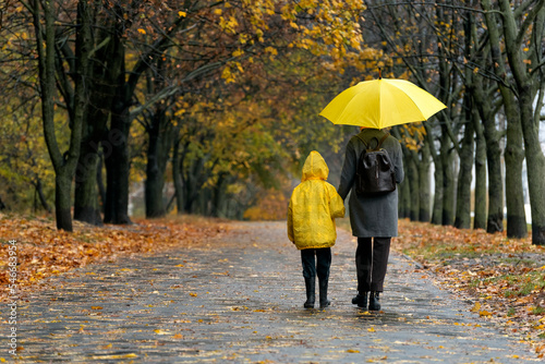Little child with woman are walking in the autumn park with large yellow umbrella and in yellow jacket . Back view.