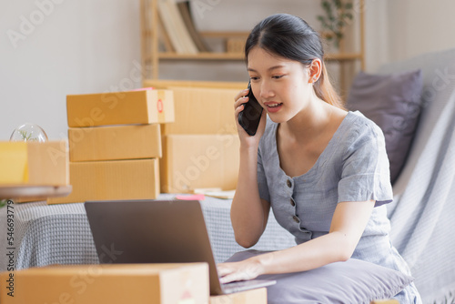 Smiling beautiful Asian woman SME entrepreneur Business owner holding cardboard boxes pack products and check customers' orders on their laptops at home, concept of SME delivery online. © David