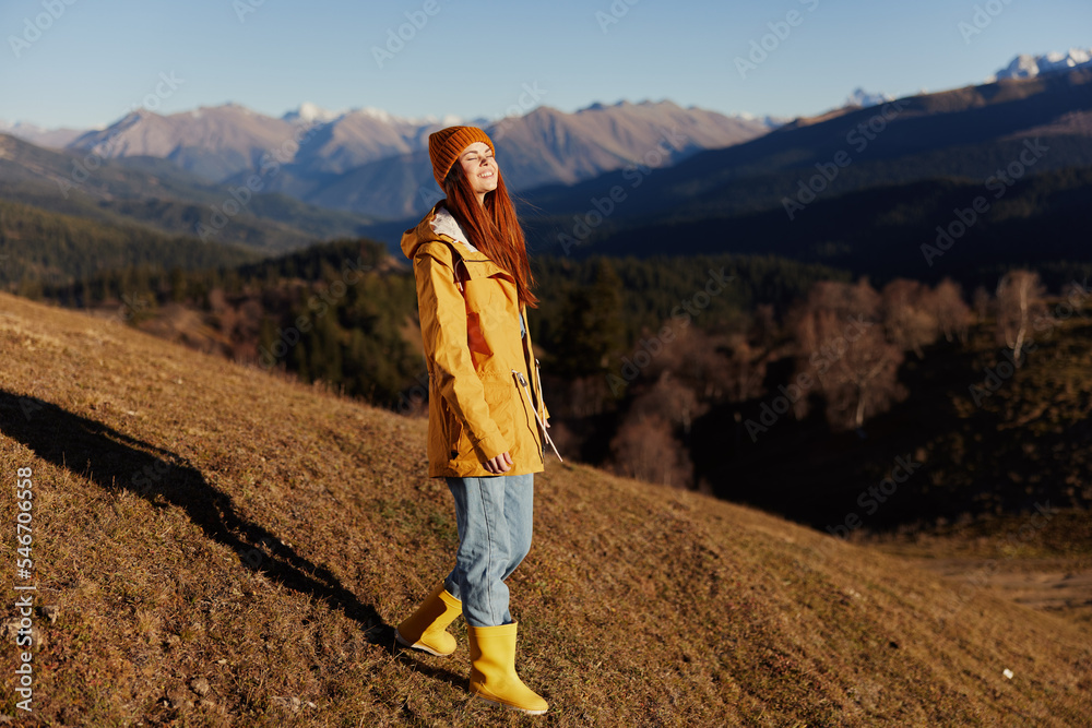 Naklejka premium Woman smile with teeth full-length walk happiness walking on the hill and looking at mountains in a yellow raincoat and jeans in the autumn happy journey in the sun sunset hiking, freedom lifestyle 