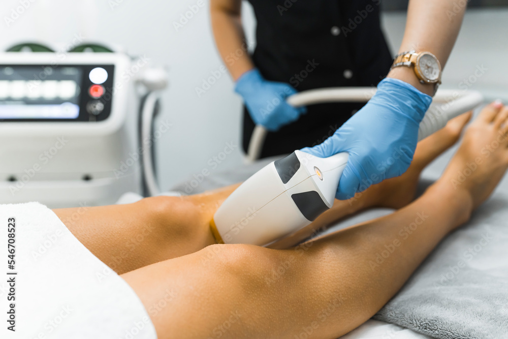 Obraz premium SPA and Beauty Salons treatment - laser hair removal from legs. Unrecognizable caucasian beautician in protective blue gloves using laser on her caucasian client. Hair removal cosmetology procedure