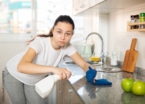 Attractive smiling young woman cleaning kitchen counter with spray bottle and rag