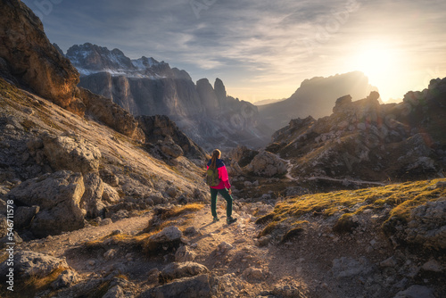 Girl with backpack on the trail in mountains at sunset in autumn. Beautiful landscape with young woman, high rocks, path, stones, orange grass, sky with clouds in fall in Dolomites, Italy. Adventure