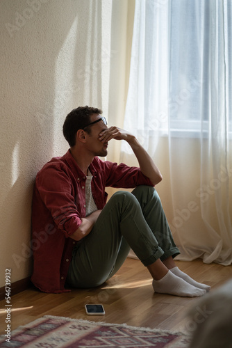 Quadro su tela Upset depressed young man sitting with closed eyes on floor at home, getting unexpected bad news over phone