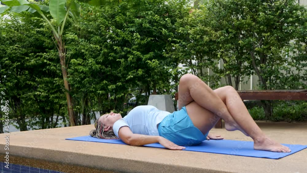 Young woman lying down on mat, crossed leg on poolside area. Does yoga excercises near home in summer time. Bush of green leaves. Gymnastic lexibility pose