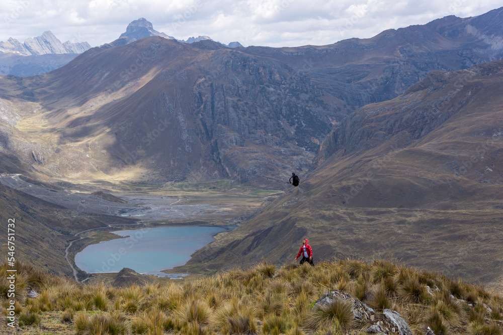 Hombre tirando hacia arriba su mochila, en una meseta o páramo, cerca a una laguna y una montaña con el cielo nublado, en los Andes Perú Sudamérica