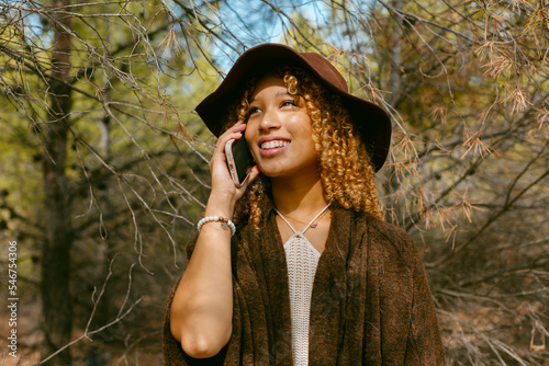 Pretty girl with hat and curly hair smiling on the phone in sunlight  photo