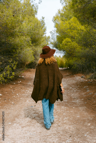 back of girl with hat and brown poncho walking in the woods 