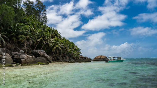 The tropical island is completely overgrown with green plants, palm trees. Picturesque boulders are piled up near the shore. A lonely boat in a calm turquoise ocean. Seychelles. Moyenne Island