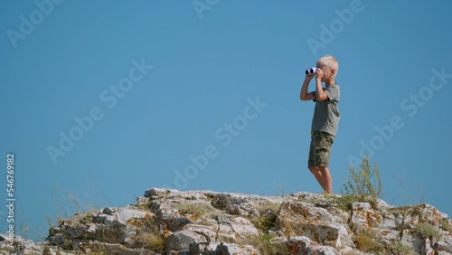 A fair-haired boy of seven years old, stands on the top of a cliff against the blue sky and looks somewhere into the distance around. He holds binoculars in his hands