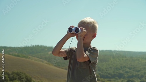 Against the backdrop of nature and the blue sky is a boy. He looks through the binoculars, which is in his hands. The boy is watching what is happening somewhere far away from him