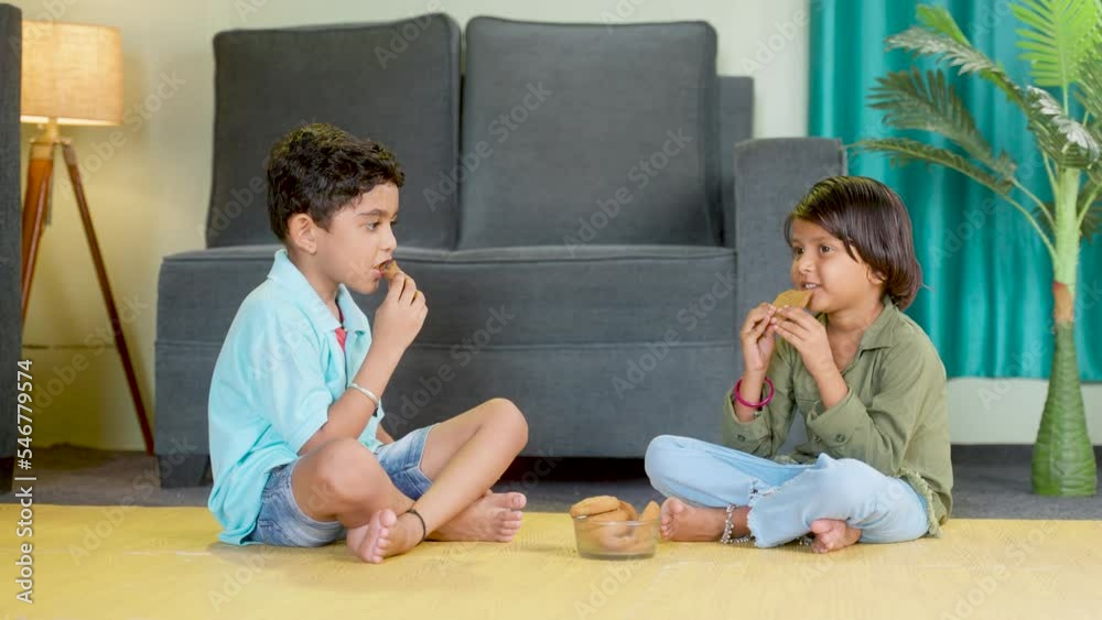 Young sibling kids having snacks or biscuit while sitting on floor at home by looking around after school - concepts of healthy eating togetherness and childhood lifestyle. Young sibling kids having snacks or biscuit while sitting on floor at home by looking around after school - concepts of healthy eating togetherness and childhood lifestyle.